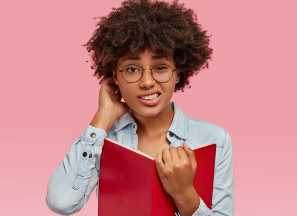 Puzzled African American woman scratches head in bewilderment, cant remember important material and give answer on question immediately, holds red book in front, dressed in fashionable jacket